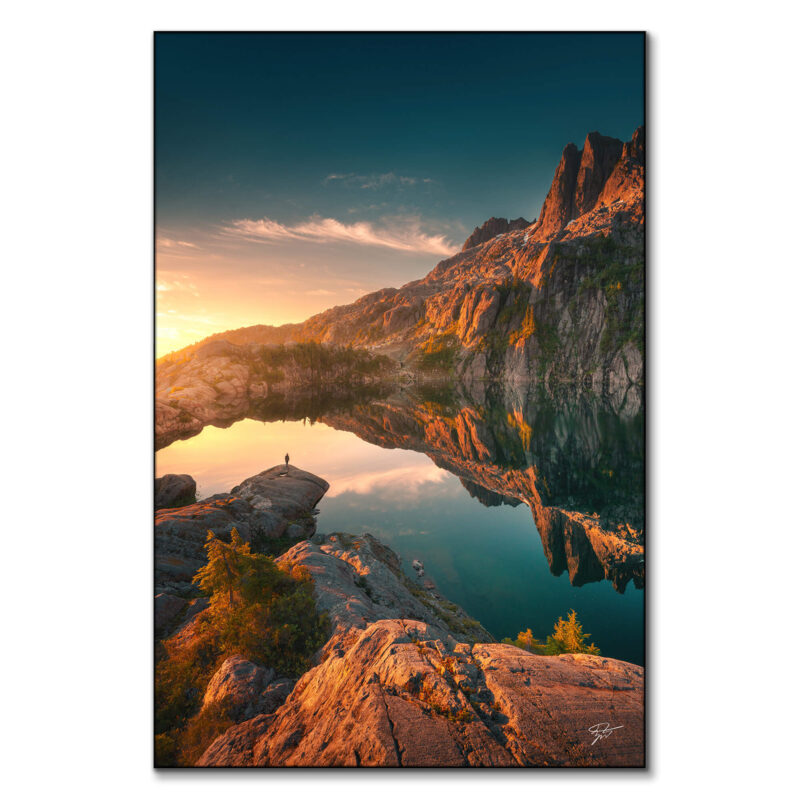 Lone figure at dawn by a pristine mountain lake in British Columbia, Canada, with mirrored reflections of rugged cliffs and peaks.