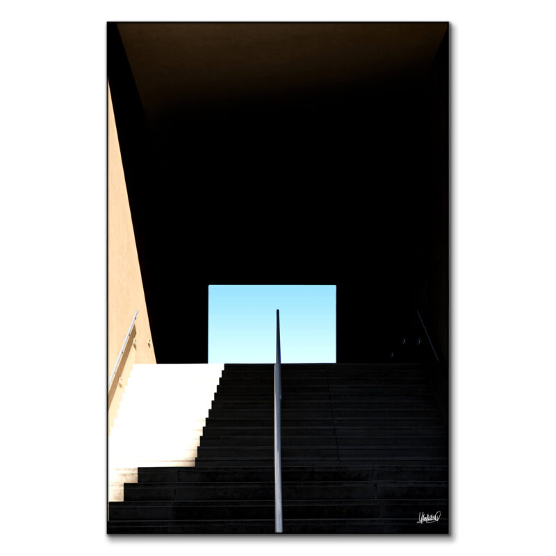 Dark staircase with a bright opening and clear blue sky, highlighting strong lines and shadows.