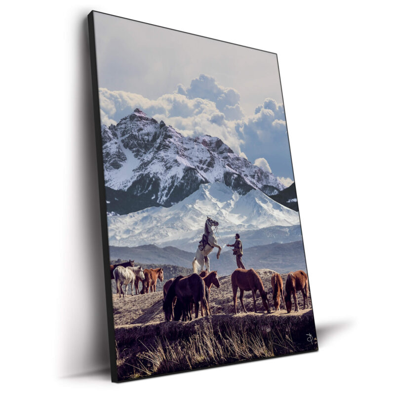 Cowboy with a horse in front of snow-capped mountains, surrounded by grazing horses in a tranquil landscape.