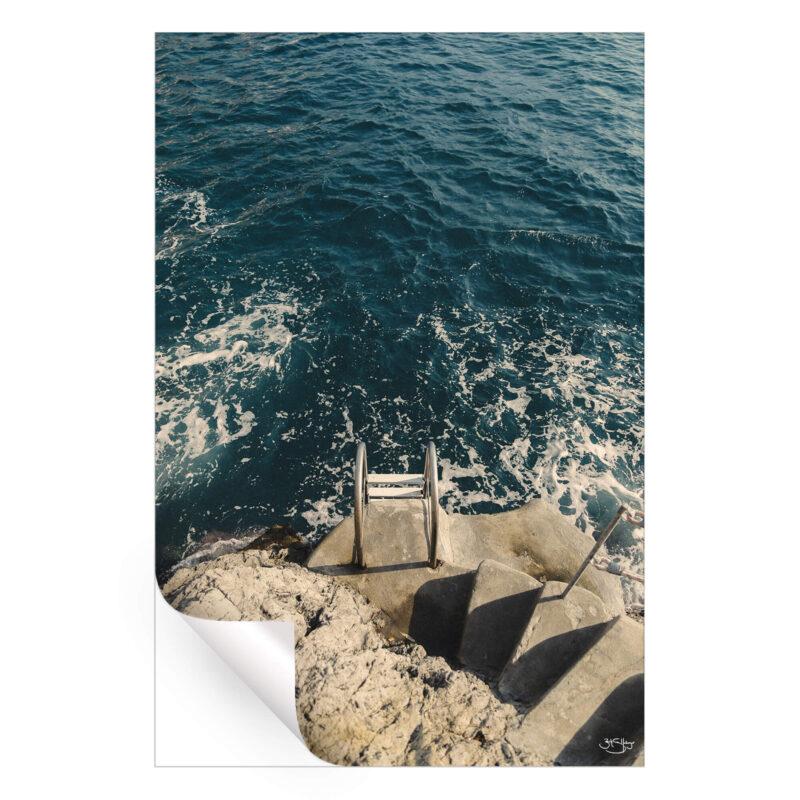 A coastal photograph featuring a stone staircase leading to a metal ladder descending into the deep blue sea with foamy waves.
