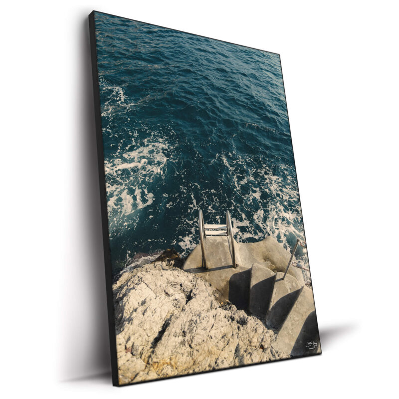 A coastal photograph featuring a stone staircase leading to a metal ladder descending into the deep blue sea with foamy waves.